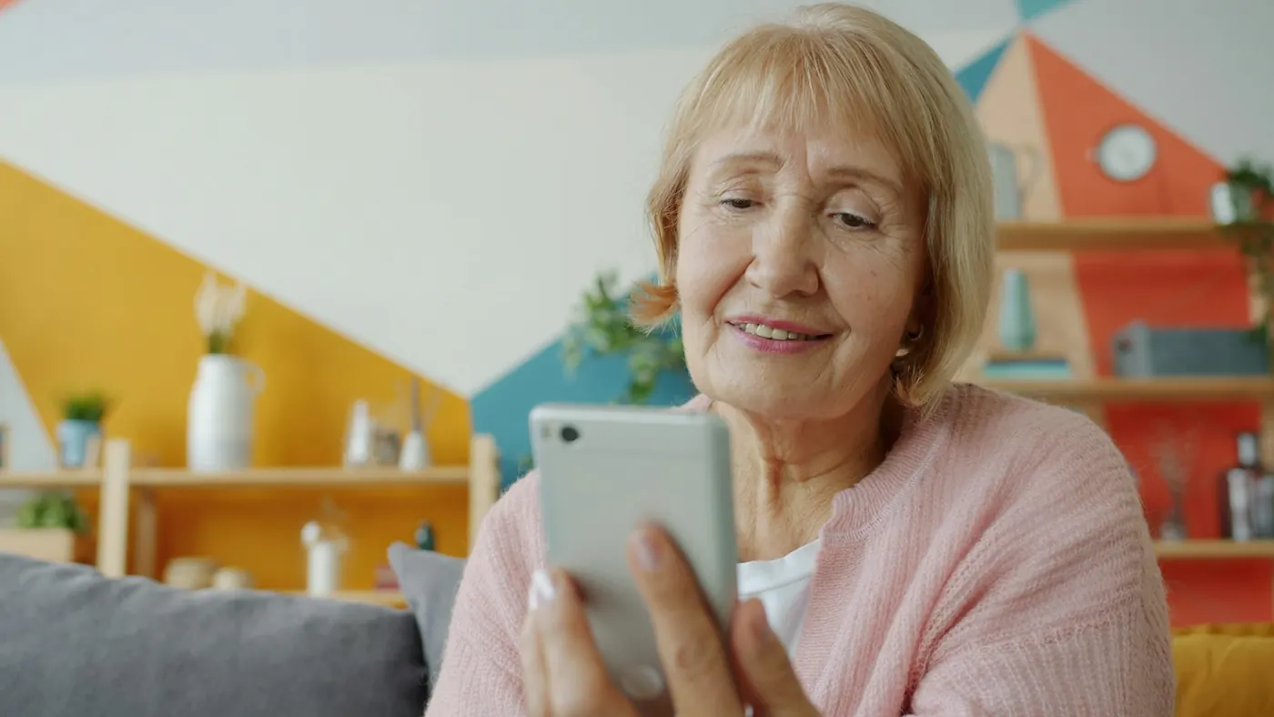Donna anziana sorridente guarda lo schermo di uno smartphone seduta sul divano di casa